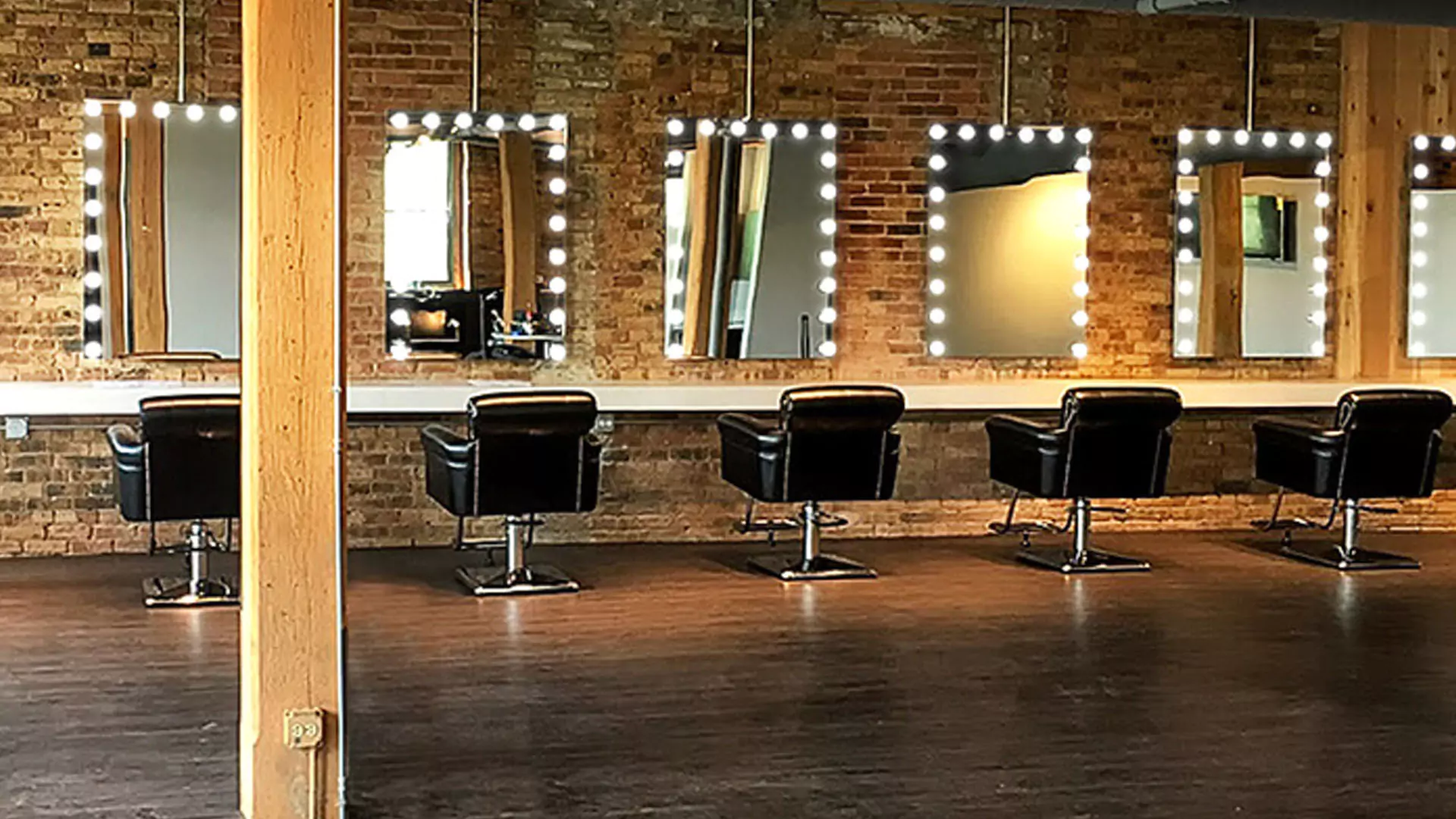 Salon with backlit hollywood lighted mirrors, brick walls, and black styling chairs in Adelaide.